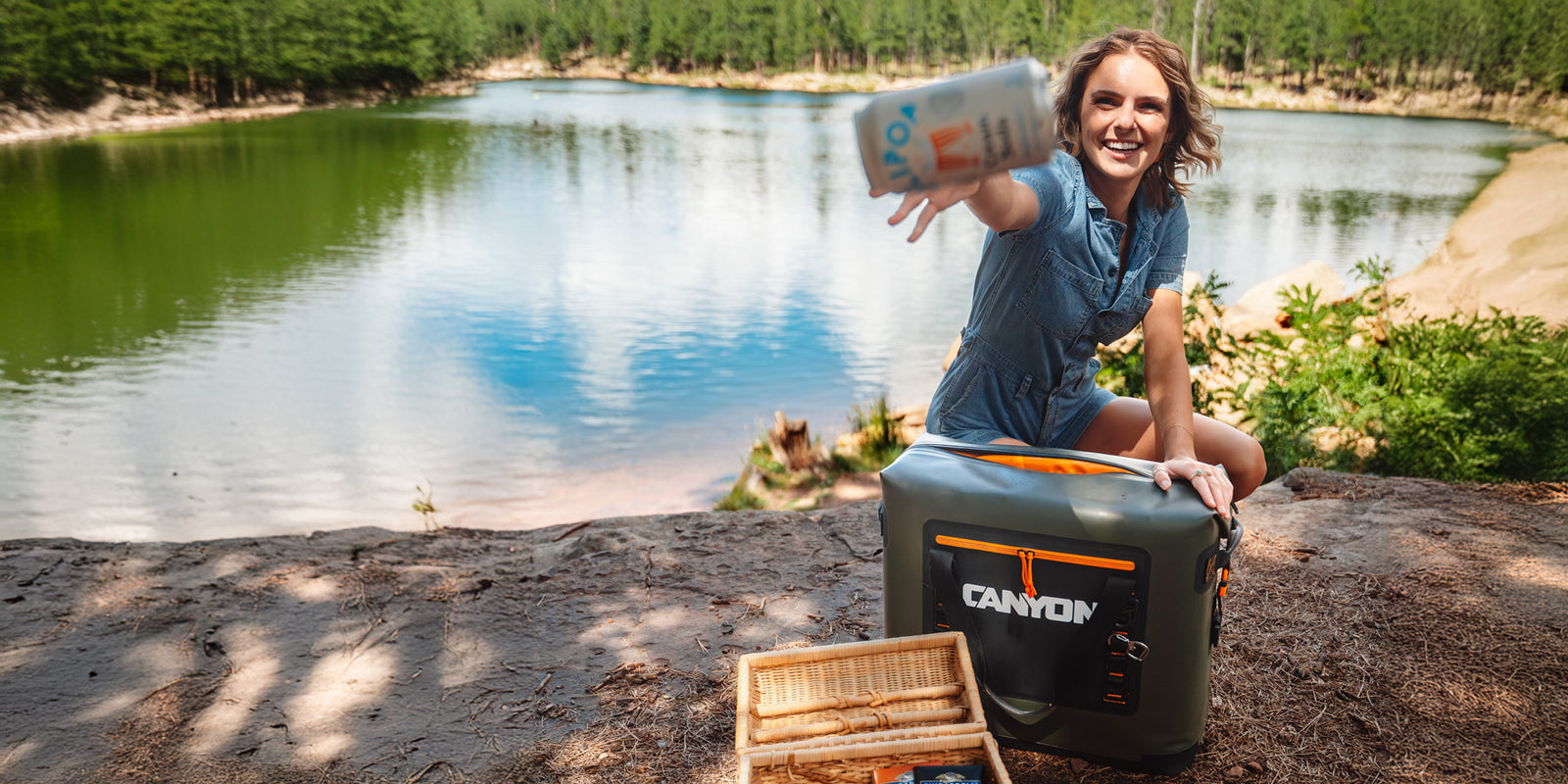 A woman throwing a soda from her Nomad 20 Soft Cooler in front of a mountain lake